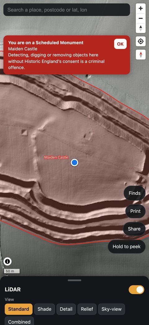 The app warning that the user is on a Scheduled Monument, Maiden Castle, where detecting, digging or removing objects without Historic England's consent is a criminal offence.